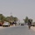 The image shows a military foot patrol moving along a paved road through what appears to be a small urban or semi-rural settlement in a Middle Eastern environment. Several soldiers in U.S. Army combat uniforms and full kit are walking in a dispersed formation along both sides of the road. Civilian vehicles, roadside shops, palm trees, and local residents are visible in the background.