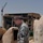 A U.S. Army soldier in ACU (Army Combat Uniform) digital camouflage stands among damaged concrete structures, looking downward at what appears to be a small opening or compartment. In the background stands a heavily damaged large building with classical architectural features, alongside a construction crane. Graffiti is visible on the concrete surface to the soldier's right.