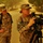 Two female U.S. Army soldiers in ACU (Army Combat Uniform) digital camouflage are operating a professional video camera on a tripod in what appears to be a dusty, hazy outdoor environment. The soldier on the right is actively reviewing or adjusting the camera while the soldier on the left stands observing. The orange-tinted haze in the background suggests a sandstorm or dust storm condition, likely in a Middle Eastern deployment location.