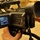A Sony professional video camera (likely a Sony DSR or similar DVCAM model) is being held and operated by a person. The flip-out LCD monitor on the side displays what appears to be footage of a military vehicle, possibly a Humvee, in an outdoor desert-like environment. The camera is equipped with a top handle, microphone mount, viewfinder, and various control buttons.