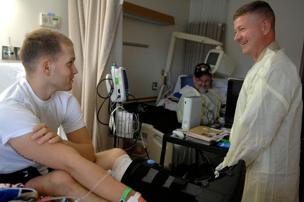 A hospital room scene showing three individuals engaged in conversation. A young male patient sits on a bed with a bandaged leg and orthopedic brace, speaking with an older man wearing a yellow medical isolation gown. A second patient is visible in the background lying in a hospital bed with a baseball cap.