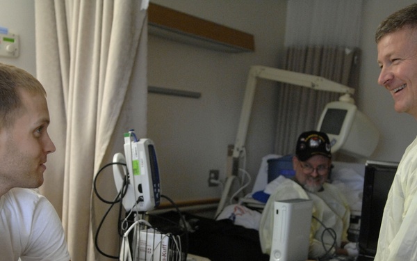 A hospital room scene showing three individuals engaged in conversation. A young male patient sits on a bed with a bandaged leg and orthopedic brace, speaking with an older man wearing a yellow medical isolation gown. A second patient is visible in the background lying in a hospital bed with a baseball cap.