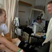 A hospital room scene showing three individuals engaged in conversation. A young male patient sits on a bed with a bandaged leg and orthopedic brace, speaking with an older man wearing a yellow medical isolation gown. A second patient is visible in the background lying in a hospital bed with a baseball cap.