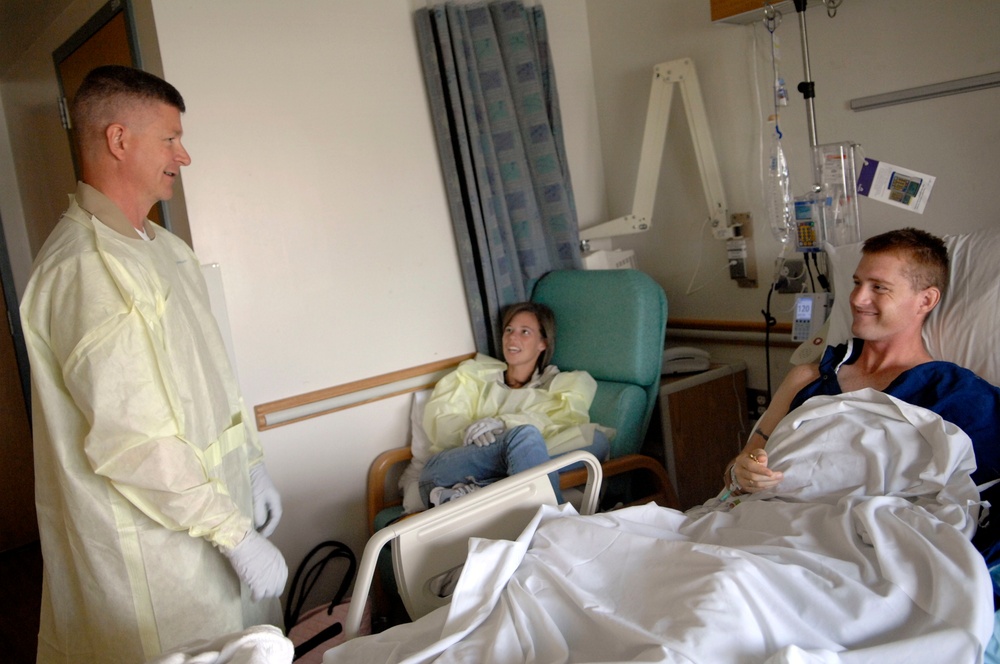 A hospital room scene showing a patient lying in a medical bed being visited by two individuals wearing yellow protective gowns and latex gloves. The patient appears to be a male in good spirits, smiling during the visit. Medical equipment including an IV stand and monitoring devices are visible in the background.