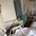 A hospital room scene showing a patient lying in a medical bed being visited by two individuals wearing yellow protective gowns and latex gloves. The patient appears to be a male in good spirits, smiling during the visit. Medical equipment including an IV stand and monitoring devices are visible in the background.