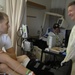 A hospital room scene showing a young male patient seated on a bed with a bandaged leg and orthopedic brace, conversing with an older man wearing a yellow medical isolation gown. A second patient is visible in the background lying in a hospital bed wearing a cap. The setting appears to be a shared military or civilian medical facility ward.