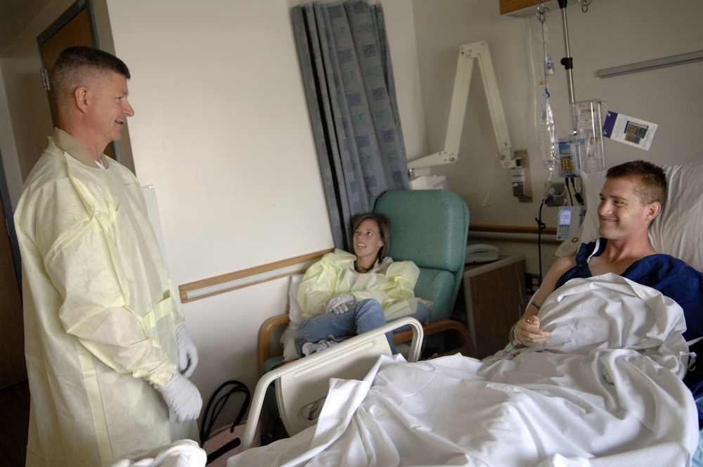 A hospital room scene showing three people engaged in conversation. A standing male visitor wearing a yellow protective gown and white latex gloves speaks with a male patient lying in a hospital bed. A female visitor, also wearing a yellow protective gown, sits in a chair between them.