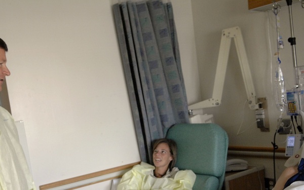 A hospital room scene showing three people engaged in conversation. A standing male visitor wearing a yellow protective gown and white latex gloves speaks with a male patient lying in a hospital bed. A female visitor, also wearing a yellow protective gown, sits in a chair between them.