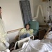 A hospital room scene showing three people engaged in conversation. A standing male visitor wearing a yellow protective gown and white latex gloves speaks with a male patient lying in a hospital bed. A female visitor, also wearing a yellow protective gown, sits in a chair between them.