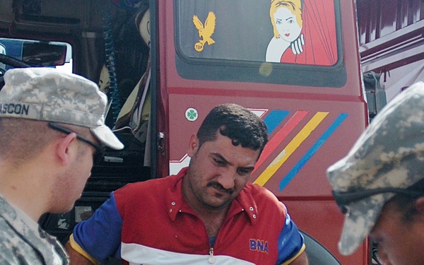 Two U.S. Army soldiers in ACU (Army Combat Uniform) camouflage are conducting what appears to be a checkpoint inspection or document check with a civilian man standing in front of a decorated red truck. One soldier is examining a document handed by the civilian while the other soldier records information on a clipboard. The setting appears to be an overseas deployment location based on the truck's decorative style and context.