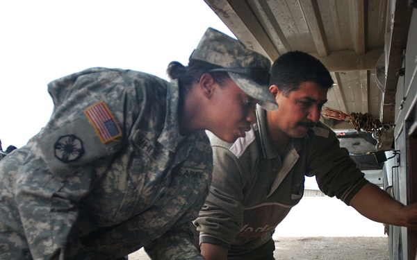 Two U.S. Army soldiers in ACU (Army Combat Uniform) camouflage pattern are conducting what appears to be a vehicle or cargo inspection alongside a civilian male. One soldier is leaning forward examining items near the underside or side compartment of a vehicle, while a second soldier observes from behind. The scene appears to take place at a checkpoint or military installation on a gravel/dirt surface.
