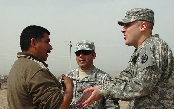 Three individuals are engaged in a conversation in an outdoor, arid environment. Two are wearing U.S. Army ACU (Army Combat Uniform) camouflage uniforms, while a third civilian man in a brown jacket faces them. The setting appears to be a forward operating base or checkpoint in a Middle Eastern location.