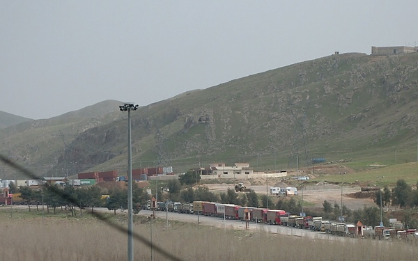 The image shows a large border crossing or truck staging area with dozens of heavy goods vehicles queued in multiple lanes. The scene is set in a hilly, semi-arid landscape with a fortified or administrative structure visible on the hilltop. Multiple company names are visible on truck trailers, suggesting international freight transport.