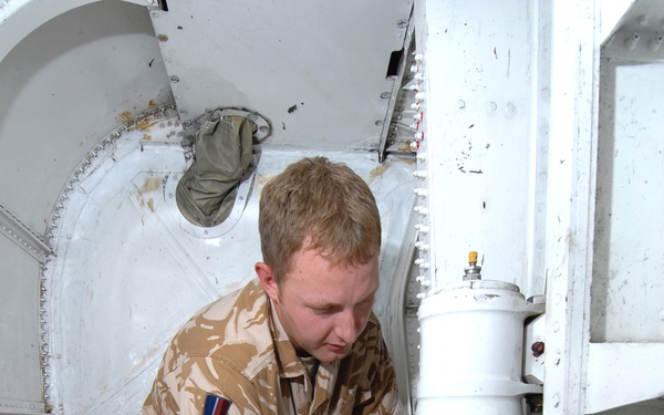 A military technician wearing British Desert DPM (Disruptive Pattern Material) camouflage uniform is working on or inspecting a cylindrical white component labeled 'C130K ONLY' inside what appears to be the fuselage or landing gear bay of a C-130K Hercules aircraft. The soldier wears a patch consistent with British Armed Forces insignia on his left sleeve. The setting appears to be a maintenance or pre-flight inspection scenario in a desert operational environment.