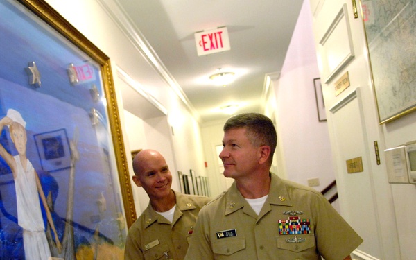 Two U.S. Navy officers in khaki service uniforms are walking through a narrow corridor, pausing to view a large framed artwork on the wall. The officer in the foreground displays multiple service ribbons and medals on his uniform, indicating senior rank. The hallway appears to be inside a formal military or government building, decorated with framed artwork and maps.