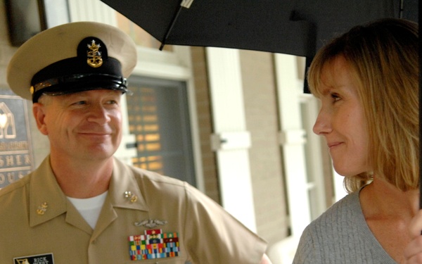 A U.S. Navy officer in tan service uniform is conversing with a civilian woman holding a black umbrella outside a Fisher House facility. The officer is smiling and has his hands on his hips, while the woman appears engaged in conversation. The setting appears to be the entrance or porch area of a Fisher House, a support facility for military families.