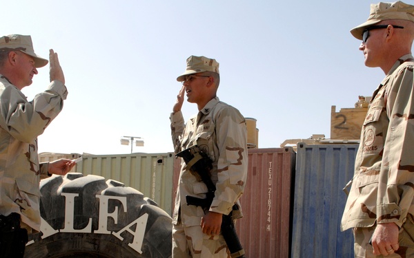 Three military personnel in desert camouflage (DCU) uniforms are gathered at a forward operating base in an arid environment. One soldier on the left appears to be gesturing or giving instructions while a center soldier salutes, and a third stands observing. A large tire display marked 'ALFA' and '74' with a diamond insignia serves as a unit marker in the foreground.