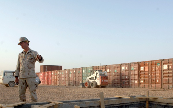 Two military personnel wearing desert camouflage uniforms and white hard hats are overseeing a construction project in an arid, sandy environment. In the foreground, a concrete foundation form is visible with wooden framing and a rebar grid laid out for a concrete pour. The background shows a row of intermodal shipping containers and a skid-steer loader (Bobcat), suggesting a forward operating base or military construction site.