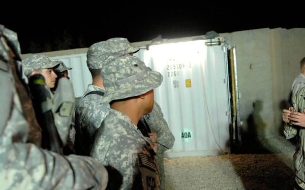 A nighttime military scene showing a US Navy officer in desert camouflage (DCU pattern) addressing a group of US Army soldiers in ACU (Army Combat Uniform) pattern. The setting appears to be a forward operating base or military compound, with shipping containers and gravel ground visible in the background. The interaction suggests a briefing or debriefing between joint service personnel.