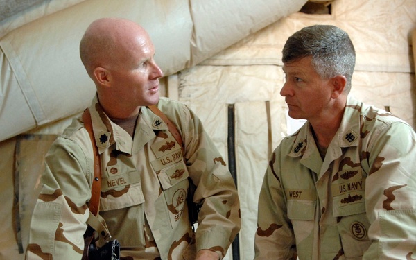 Two U.S. Navy personnel in desert camouflage (DCU) uniforms are engaged in a briefing or planning discussion inside a military tent structure. They are leaning over a table examining what appears to be a chart or map. Name tapes visible on their uniforms read 'NEVEL' (left) and 'WEST' (right).