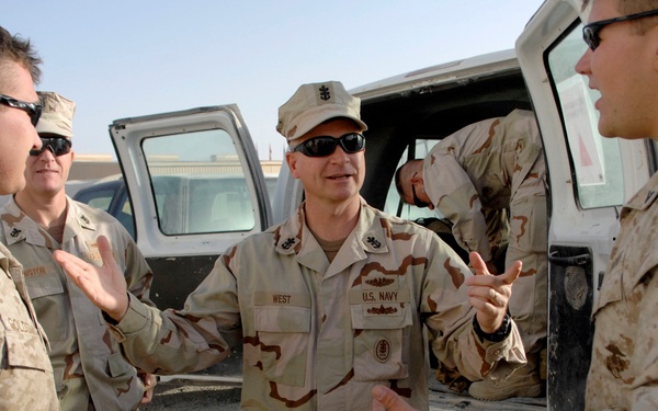 Several military personnel in desert camouflage uniforms are engaged in conversation near a military vehicle. The central figure is wearing a U.S. Navy uniform with the name tape 'WEST' visible, while others appear to be wearing USMC MARPAT desert uniforms. The setting appears to be an outdoor military installation in an arid environment.