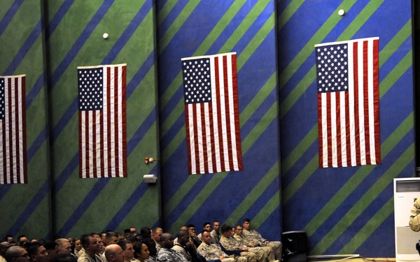 A military briefing or address is taking place inside a large indoor facility, likely a hangar or assembly hall. A single soldier in desert camouflage (ACU/DCU) uniform is speaking into a microphone before a large seated audience of military personnel. Four American flags are displayed prominently on the decorated wall behind the audience.