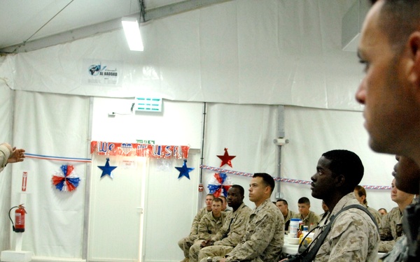 A senior military officer in desert camouflage uniform is addressing a group of seated soldiers in what appears to be a tent or temporary facility decorated with American patriotic decorations. The soldiers, wearing various camouflage uniforms and carrying rifles, are attentively listening to the briefing or address. The setting suggests a forward operating base or military installation, likely in a Middle Eastern deployment.