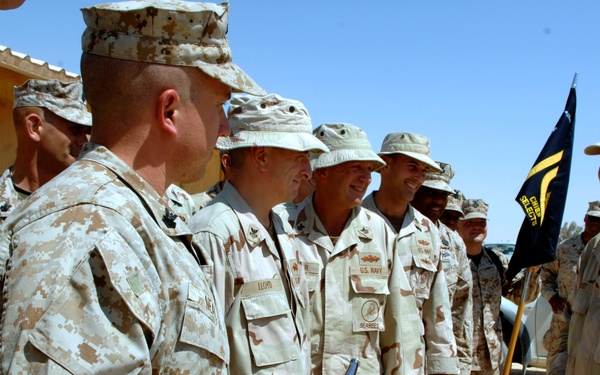 A group of military personnel in desert camouflage uniforms are gathered outdoors in what appears to be a forward operating base or military installation in an arid environment. A senior officer on the right, wearing U.S. Navy insignia, appears to be greeting or addressing a formation of troops. The scene suggests an official visit, inspection, or commendation ceremony.