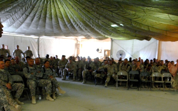 A military briefing or town hall meeting is taking place inside a large tent or temporary structure. A senior officer in desert camouflage (DCU pattern) is addressing a large group of seated soldiers using a handheld microphone, while another soldier in ACU pattern stands nearby. The audience consists of dozens of uniformed military personnel seated in folding chairs.
