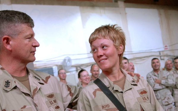 Two U.S. Navy personnel in desert camouflage (DCU) uniforms are engaged in conversation in what appears to be a military facility or warehouse. The individual on the left has the name tape 'WEST' and the one on the right has 'BOYD', with Boyd carrying a slung rifle. A group of additional military personnel are visible in the background.