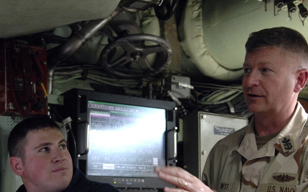 Two U.S. Navy personnel are inside what appears to be a submarine or naval vessel control room. One sailor in a dark navy uniform is seated at a workstation with a monitor displaying data, while a senior officer in desert camouflage (DCU) uniform gestures while speaking. The environment shows dense equipment, piping, and control systems typical of a submarine interior.