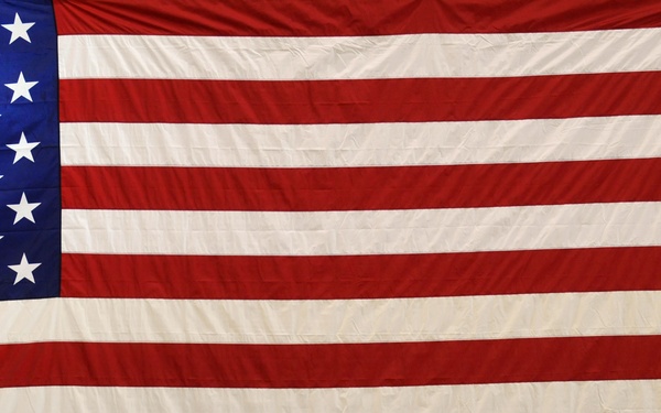 A large American flag is displayed on a wall, dominating the frame. In the lower right corner, a military officer in tan/khaki dress uniform stands at a podium with a microphone, appearing to deliver a speech or address. The setting appears to be an indoor gymnasium or assembly hall.