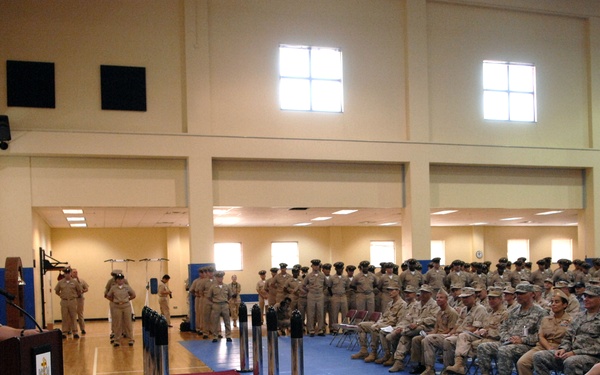 A military ceremony or formation is taking place inside a gymnasium or indoor sports facility. A uniformed officer is speaking at a wooden podium equipped with a microphone, addressing a large audience of seated and standing military personnel in tan/khaki uniforms. The setting appears to be a formal military event such as a change of command, promotion ceremony, or unit briefing.