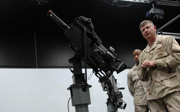 A U.S. Navy senior enlisted or officer personnel is conducting a briefing or training session for a group of sailors in a simulator or training facility. The instructor stands on an elevated platform next to what appears to be a weapons system simulator mount, addressing uniformed personnel standing in formation. All individuals are wearing desert camouflage (DCU - Desert Camouflage Uniform) pattern uniforms.
