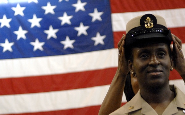 A U.S. Navy service member in the foreground is placing or adjusting a chief petty officer combination cover (cap) on their head during what appears to be a promotion or pinning ceremony. The individual is wearing a khaki service uniform adorned with multiple ribbons, warfare devices, and collar insignia. Two other Navy personnel in similar uniforms stand in the background before a large American flag.