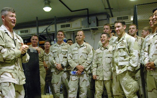 A senior military officer in desert camouflage (DCU pattern) addresses a group of approximately 15-20 service members in an indoor facility. The group appears engaged and smiling, suggesting an informal briefing or morale visit. Some personnel have 'U.S. NAVY' name tapes visible on their uniforms.