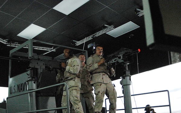 Military personnel in desert camouflage uniforms are operating what appears to be a U.S. Navy combat simulation training platform. The platform, labeled 'U.S. NAVY,' is elevated and equipped with mounted weapons, surrounded by projection screens simulating a maritime environment. Several sailors/soldiers are engaged in the simulation exercise while others observe from the floor level.
