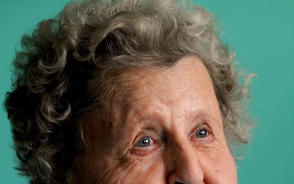 A formal portrait photograph of an elderly woman with curly gray hair, photographed against a teal/turquoise background. She is wearing a light blue cardigan over a white collared shirt and displays a medallion pendant on a silver chain. Her expression is engaged and slightly open-mouthed, as if mid-conversation.