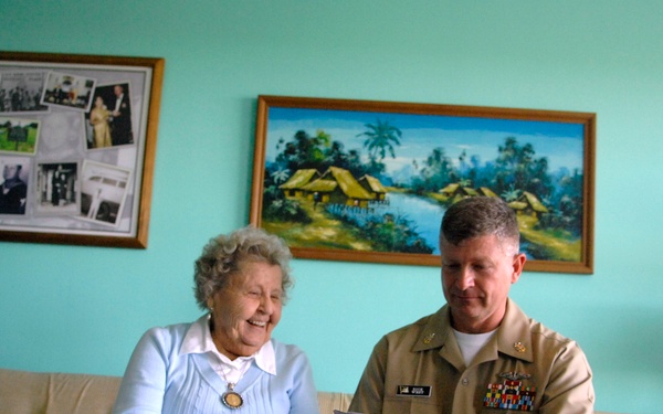 An elderly woman and a male military officer in tan/khaki uniform are seated together on a beige sofa in a home living room. The officer is holding and reviewing what appears to be a card or document while both individuals appear engaged and smiling. The setting suggests a personal or ceremonial visit.