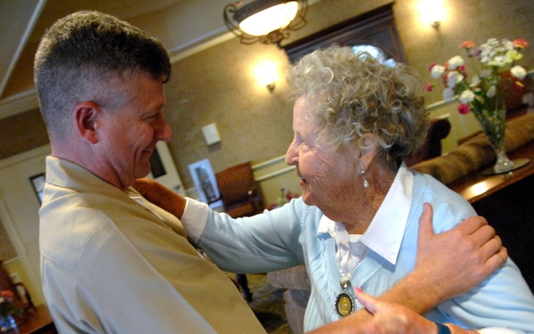 A man in a tan/khaki military-style uniform is warmly greeting an elderly woman with curly white hair in what appears to be a hotel lobby or assisted living facility common room. The two are embracing and smiling at each other, suggesting a joyful reunion. The setting is well-furnished with warm lighting, floral arrangements, and patterned carpeting.