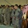 A formal ceremony showing a mixed group of military personnel and civilian dignitaries standing in a row with heads bowed, suggesting a moment of prayer or silence. The group includes U.S. Army soldiers in ACU (Army Combat Uniform) pattern, Marines in MARPAT desert camouflage, and individuals in traditional Arab Gulf attire (thobe and keffiyeh). The setting appears to be an indoor venue such as a hangar or large hall.