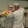 Two U.S. Marines in MARPAT desert camouflage uniforms are performing a military colors ceremony, appearing to case or uncase a unit guidon or colors with red and yellow fringe on a wooden staff. Several other military personnel are visible in the background, including individuals in desert tri-color camouflage uniforms consistent with Iraqi military dress. The ceremony appears to be taking place inside a large building or hangar.