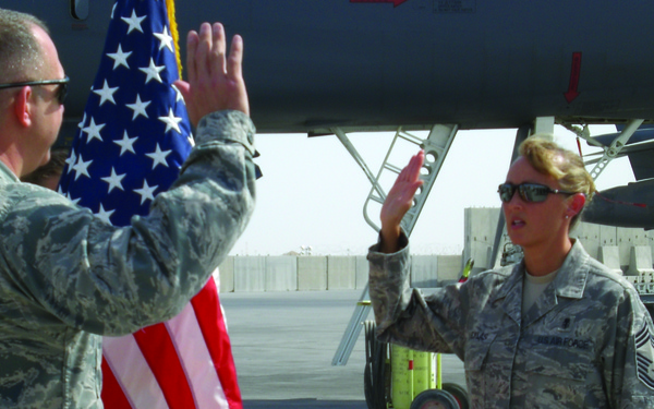 Two U.S. Air Force personnel in ABU (Airman Battle Uniform) camouflage are conducting a military oath ceremony on an airfield tarmac. Both individuals have their right hands raised, with an American flag positioned between them. The setting appears to be an overseas or deployed location based on the arid environment and concrete blast barriers visible in the background.