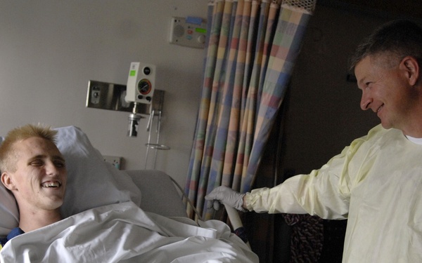 A young male patient with blonde/red hair is lying in a hospital bed, smiling and engaged in conversation. A middle-aged man wearing a yellow protective isolation gown and gloves stands beside the bed, leaning in and smiling back. The setting is a hospital room with medical equipment visible in the background.