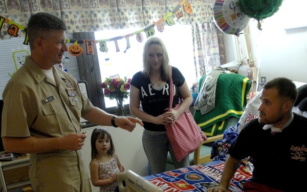 A military officer in tan/khaki uniform is visiting a patient in what appears to be a military or veterans hospital room decorated for Halloween. The patient, a young man seated on a hospital bed covered with a USMC-themed blanket, is accompanied by a young woman and a small child. The officer appears to be presenting or handing something to the patient during the visit.