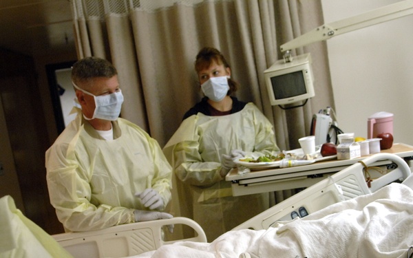 A male patient is lying in a hospital bed with an IV line and identification wristband on his wrist. Three individuals wearing yellow isolation gowns and surgical masks are present in the room, suggesting infection control precautions are in place. A meal tray with food items is visible on a bedside table, indicating the patient is receiving routine care.