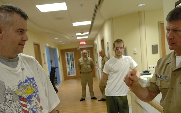 A military officer in tan/khaki dress uniform with medals and ribbons is conversing with a man in a white graphic t-shirt wearing a yellow wristband, likely in a medical facility hallway. In the background, two additional individuals in military attire are visible, one of whom appears to be a patient in a white t-shirt and green pants. The setting appears to be a military hospital or rehabilitation center.