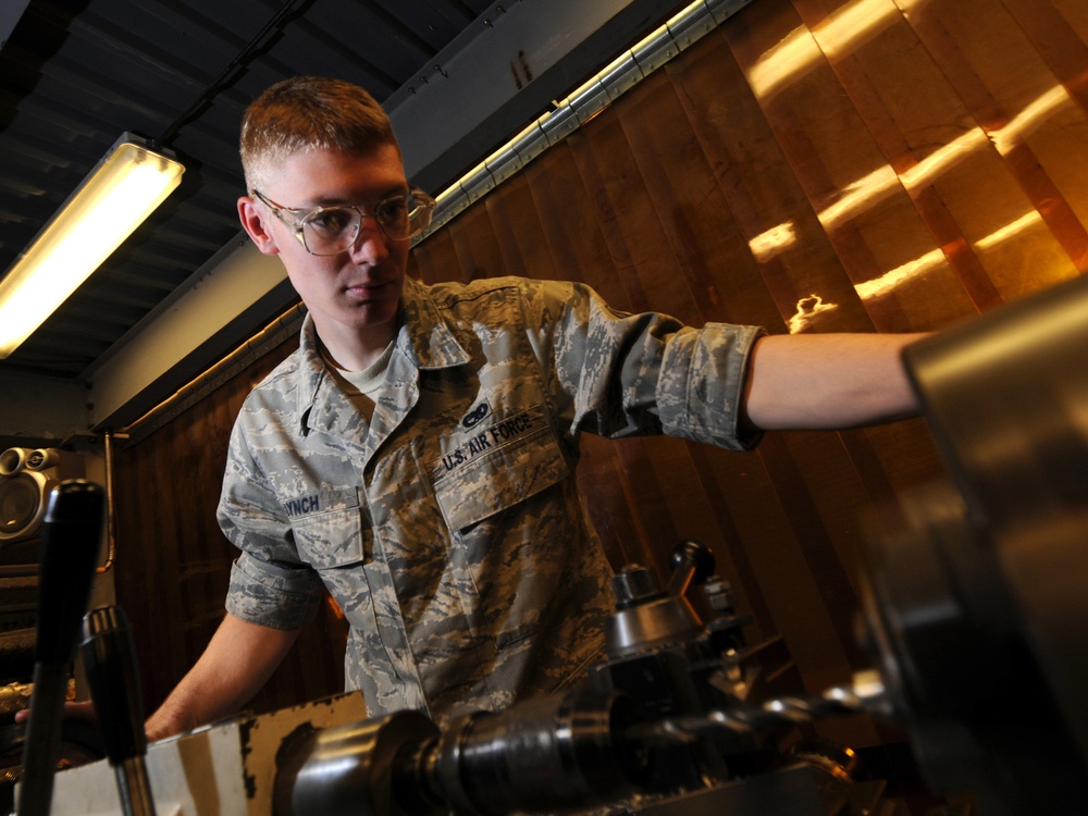 A U.S. Air Force servicemember wearing ABU (Airman Battle Uniform) and safety glasses is operating or adjusting machinery in what appears to be a maintenance or machine shop environment. The individual's name tape reads 'LYNCH' and the branch tape reads 'U.S. AIR FORCE'. The photo is taken from a low angle, giving a dynamic perspective of the work being performed.