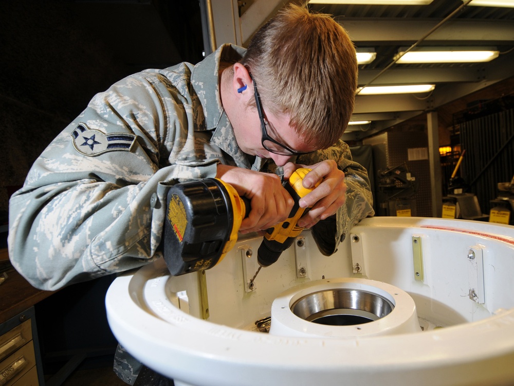 A U.S. Air Force enlisted member in ABU (Airman Battle Uniform) is using a yellow DeWalt cordless drill to perform maintenance or assembly work on a white circular equipment housing. The airman is wearing safety glasses and blue foam ear protection, indicating compliance with workshop safety protocols. The setting appears to be a military maintenance facility or hangar bay with fluorescent overhead lighting.