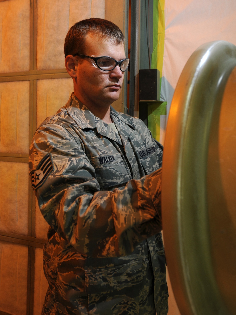A U.S. Air Force servicemember named Walker, wearing ABU (Airman Battle Uniform) camouflage and safety glasses, is focused on inspecting or working on a large olive-drab colored object, likely munitions or ordnance. The rank insignia on the left sleeve indicates the rank of Senior Airman. The setting appears to be an indoor maintenance or storage facility with warm orange lighting.